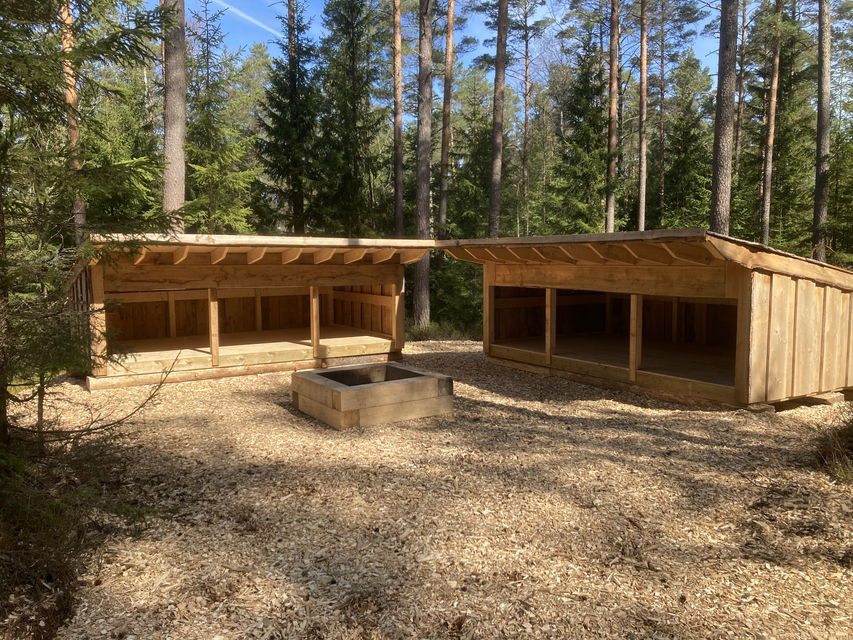 Två vindskydd och en grillplats med skog i bakgrunden