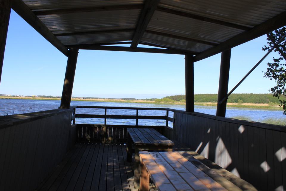 På fågeltornets andra våning står två bord och en lång bänk. Det är höga skyddsräcken runt och ett tak över.