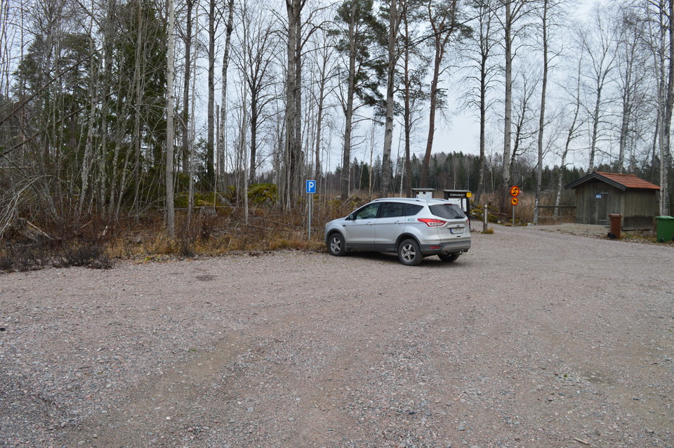 En bil står på en rymlig grusad parkeringsplats. I bakgrunden står en torrtoalett.