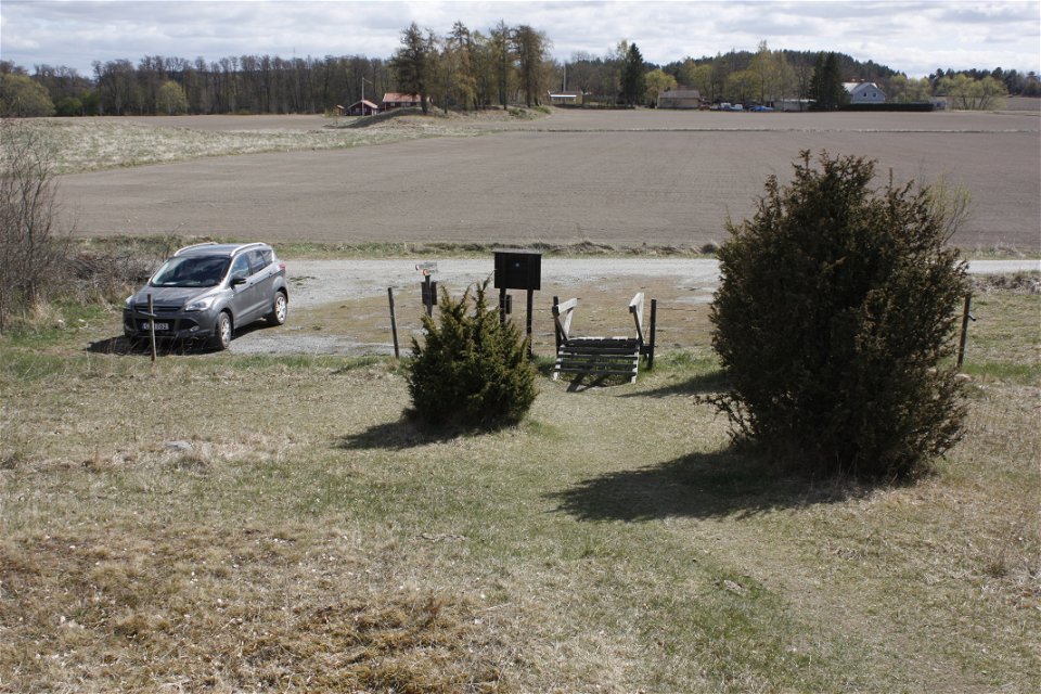En bil står parkerad på en grusad yta vid en grusväg. Framför parkeringen står en informationsskylt och ingång in till en beteshage.