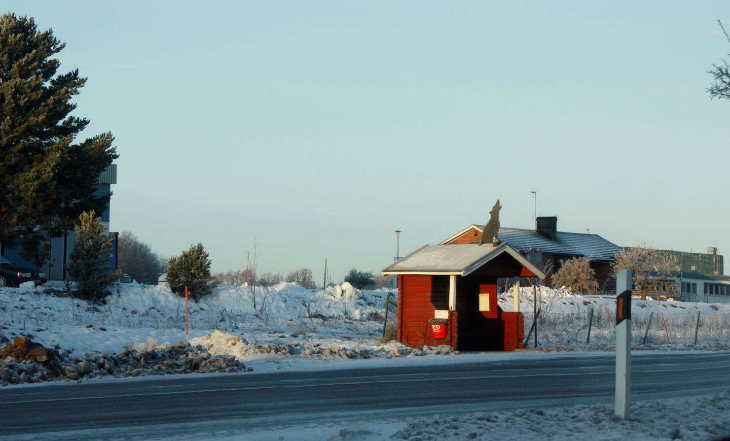 En busskur står vid en bilväg. Bakom kuren finns flera byggnader och det ligger snö på marken.
