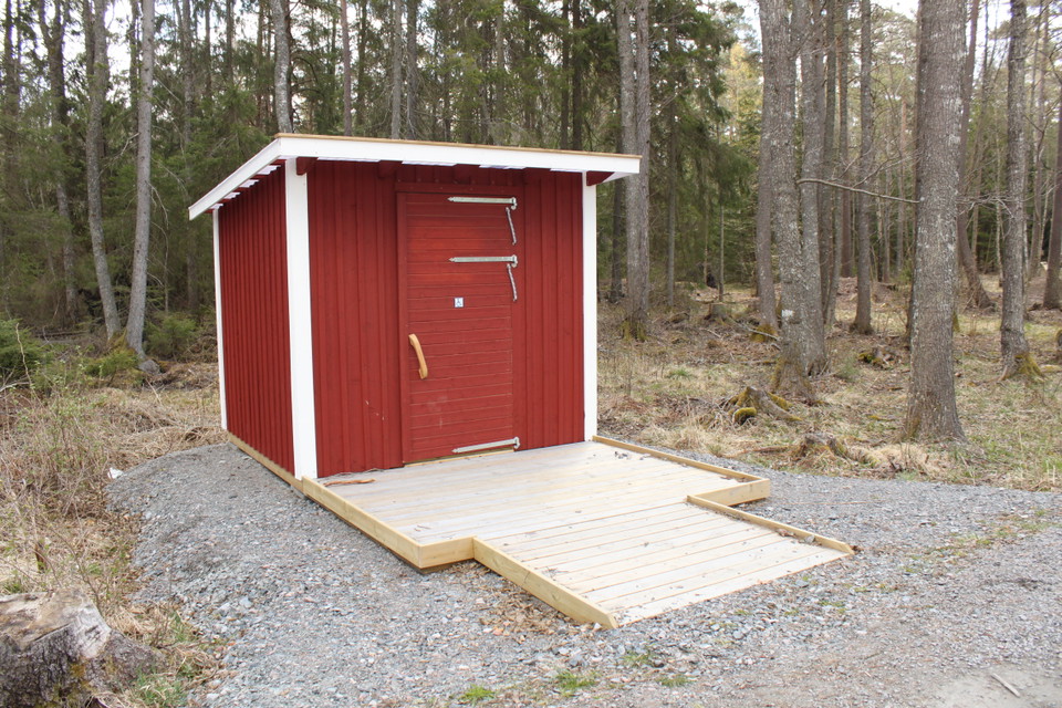 En tillgänglighetsanpassad torrtoalett står på en plan grusad yta med skog i bakgrunden. En bred, låg träramp leder upp till toalettdörren.