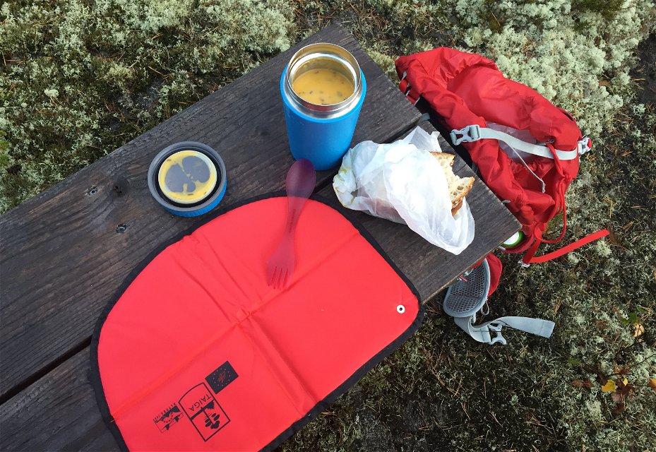 Öppen mattermos, smörgås och sittunderlag på bänk. En röd ryggsäck står på marken intill bänken.