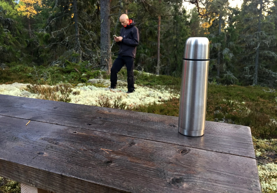 En termos på en bänk. En man står i bakgrunden och tittar på sin mobiltelefon.