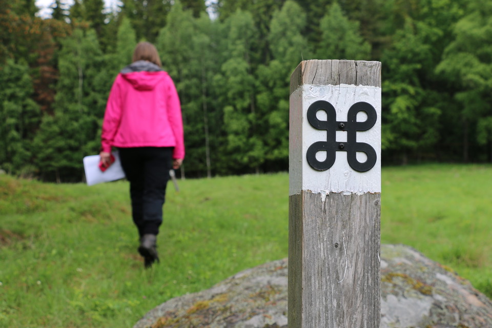 Vitmålad stolpe med en svart kringla som är symbolen för fornminne. På gräsmark går en person bortåt mot skogen.