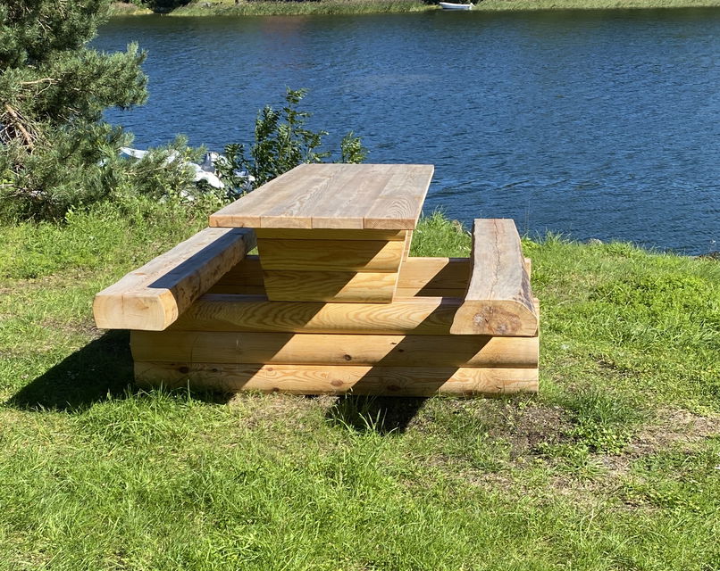 Ett träbord med fasta bänkar står på en gräsmatta. Vatten syns i bakgrunden.
