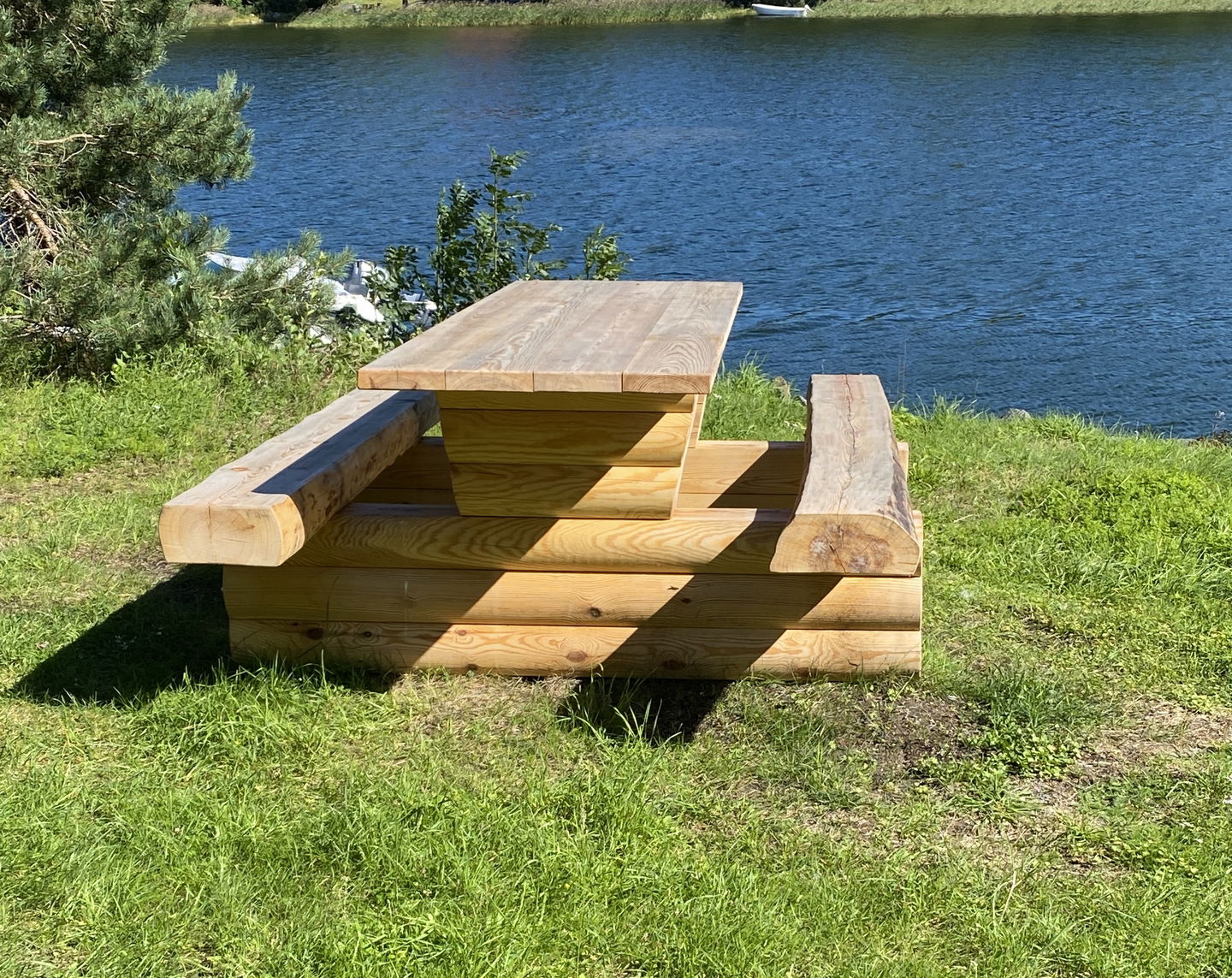 Ett träbord med fasta bänkar står på en gräsmatta. Vatten syns i bakgrunden.