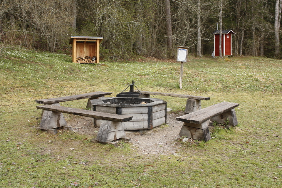 En grillplats med fyra sittbänkar runtom står på en stor öppen gräsyta. I bakgrunden står ett vedförråd och ett torrdass.