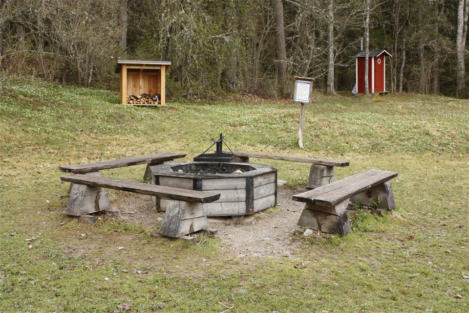 En grillplats med fyra sittbänkar runtom står på en stor öppen gräsyta. I bakgrunden står ett vedförråd och ett torrdass.