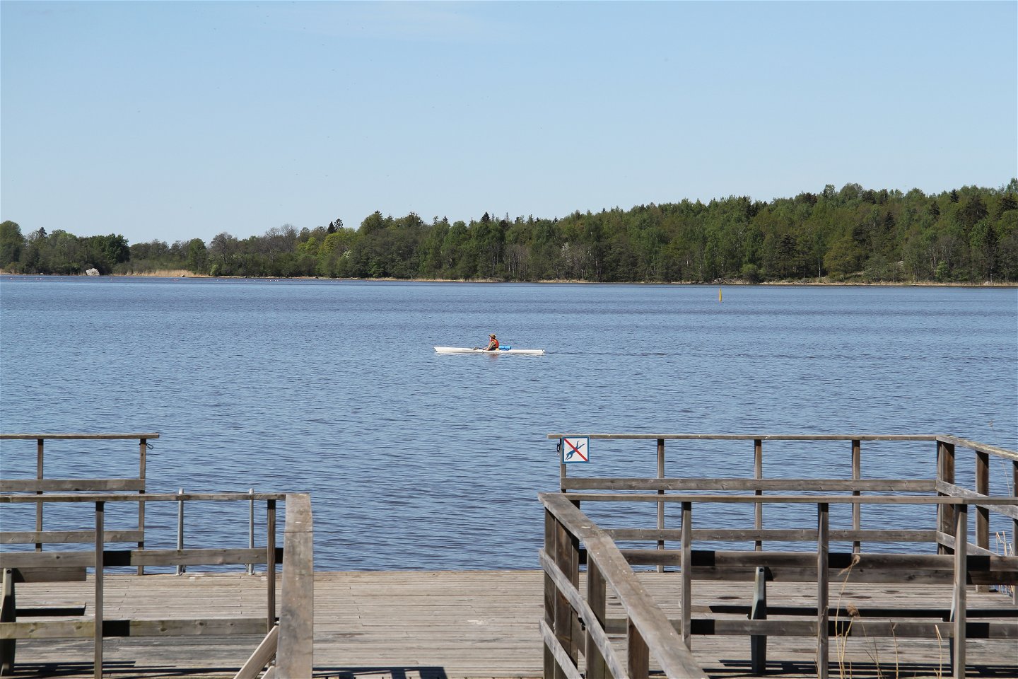 Brygga med träräcke och en skylt som visar att dykning är förbjuden. På sjön syns en person paddla kajak.