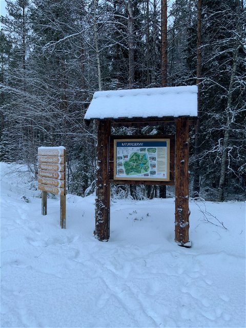 En informationstavla som sitter på ett ställ med tak. Bredvid tavlan finns stigvisare med 6 destinationer i Florarna. Det är snö på marken. 