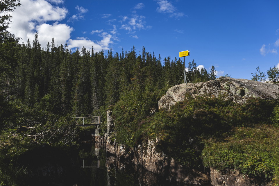 En träbro över ett vatten och en gul skylt på en stenklippa