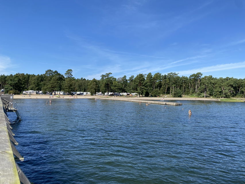 Sandstrand med brygga. Camping och tallskog i bakgrunden