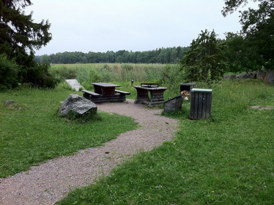 En upphöjd eldstad med grillgaller står bredvid ett bord med två bänkar. Soptunnor och vedförråd står intill. Platsen ligger nära vattnet.