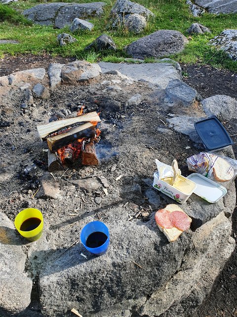 En grillplats med en liten eld som brinner, runt om står smörgåsar, smörpaket, muggar med dryck.