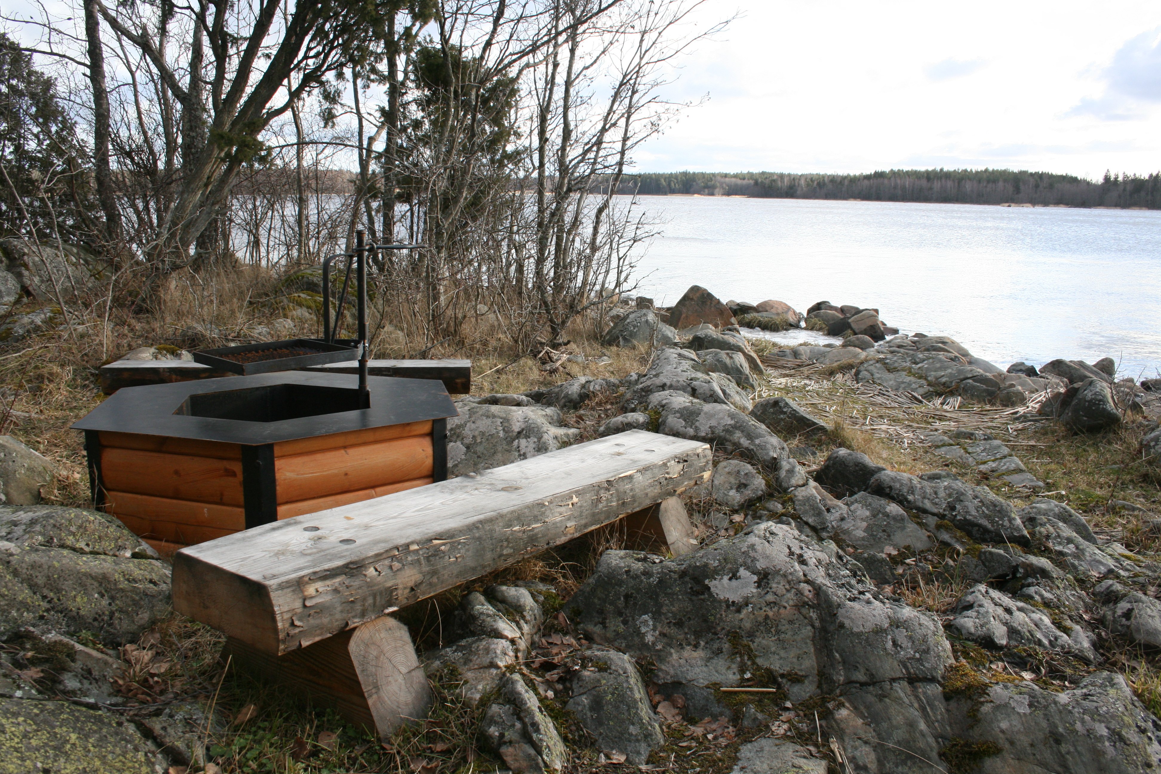 En låg grillplats med två sittbänkar bredvid står bland stora stenar vid havet.