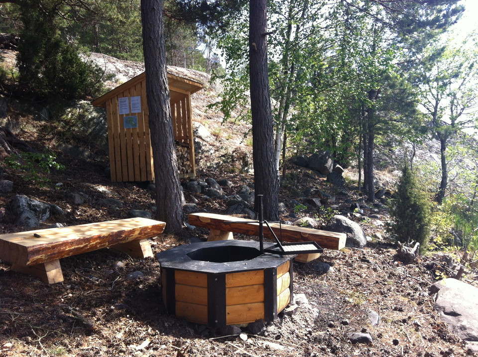 En eldstad med två sittbänkar bredvid. I bakgrunden syns ett litet vedförråd. 