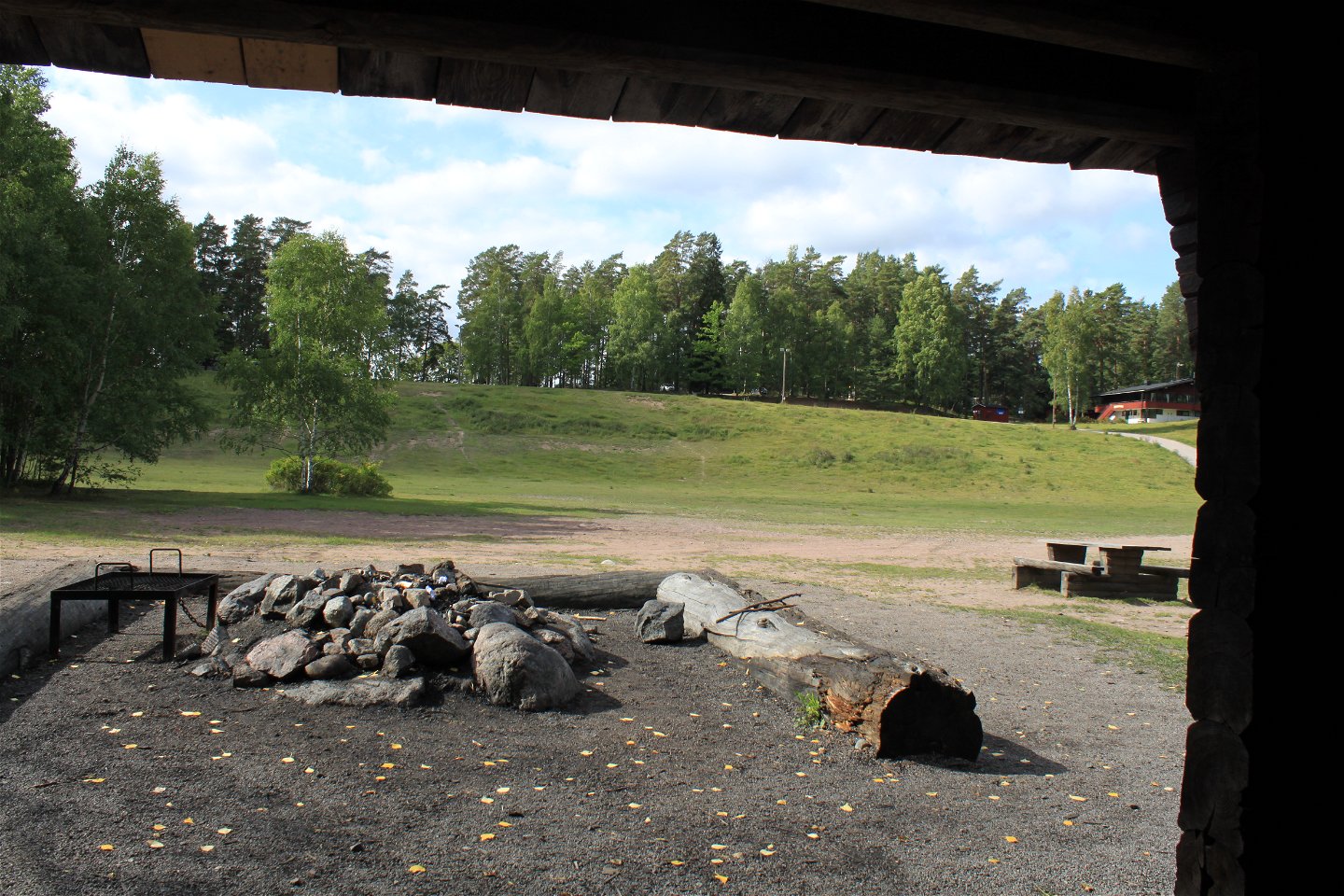 Från vindskyddets öppning kan man se grillplats med låga stockar runtom. I bakgrunden syns Sunnerstaåsen och många träd.