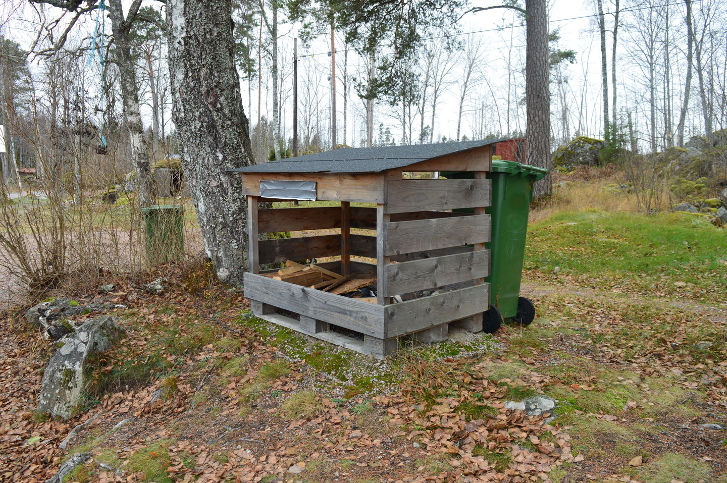 Ett litet öppet vedförråd med tak över står på ojämn mark. Bakom vedförrådet står en soptunna.