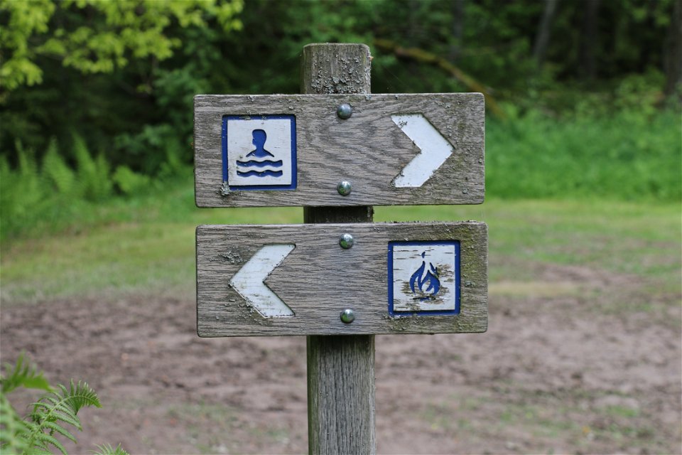 Två träskyltar som visar vägen till rastplats och badplats.