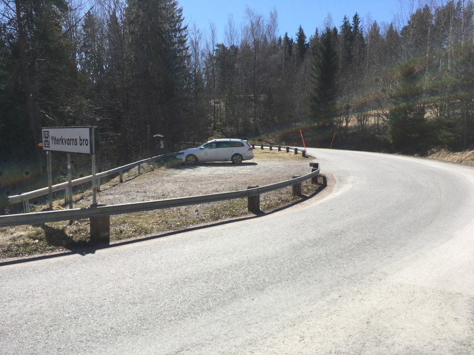 En bil står i en grusad parkeringsplats vid sidan av en bilväg. På parkeringen står en skylt med texten "Ytterkvarns bro".