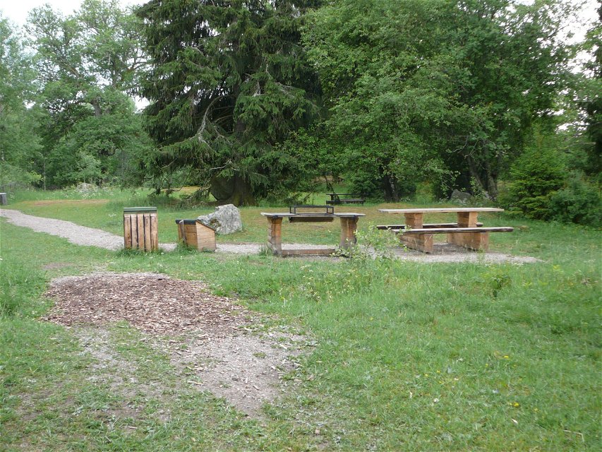 Ett grillbord står på en grusad yta. Bredvid finns bänkbord och en soptunna och i bakgrunden är det skog.