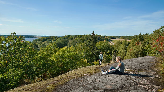 På ett högt berg sitter ett barn och tittar ut över skog och vatten. Ett annat barn står längre bort i bilden.