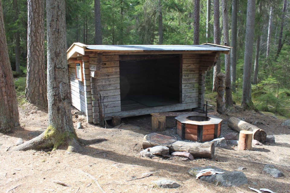 I en skogsglänta står ett vindskydd med en grillplats och sittbänkar framför.