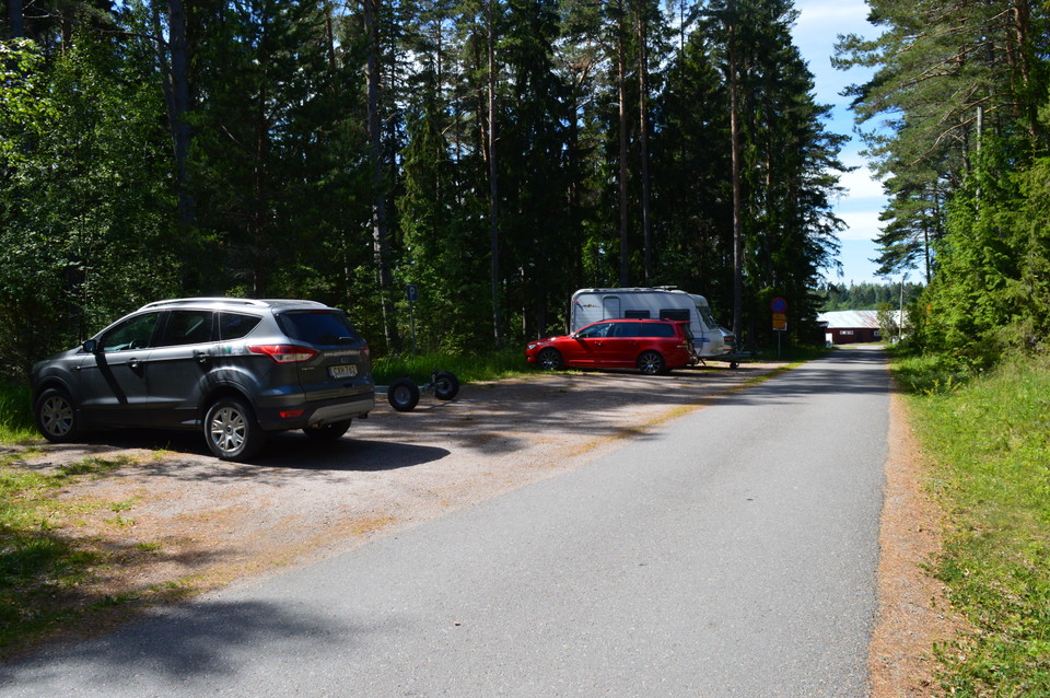 Längs en smal asfalterad väg står flera bilar och ett släp parkerade på en avlång parkering av hårdpackat grus. Runtom parkeringen växer det många träd.