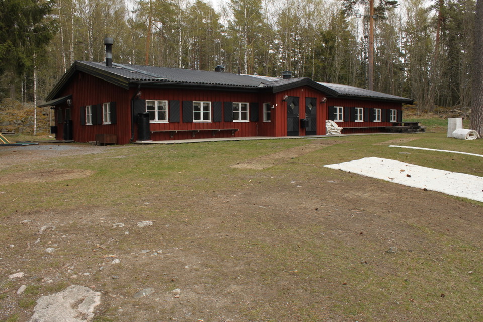 På en öppen yta står en lång husbyggnad med sittbänkar utanför. Bakom stugan är det skog.