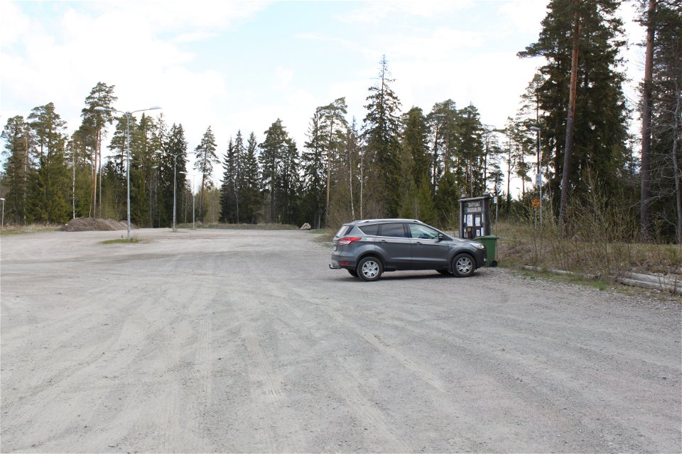 En bil står på en mycket stor grusad parkeringsplats vid en skog. På parkeringen finns en informationstavla och gatubelysning.