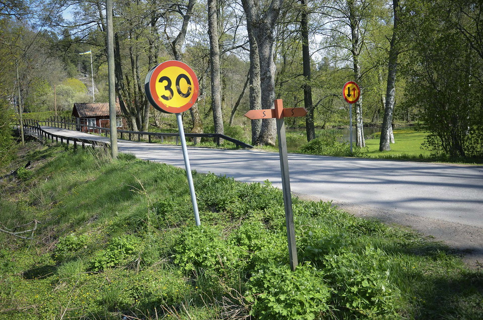 Liten asfaltsväg med grönsak runt om, en Sörmlandsledenskylt och vägskyltar.