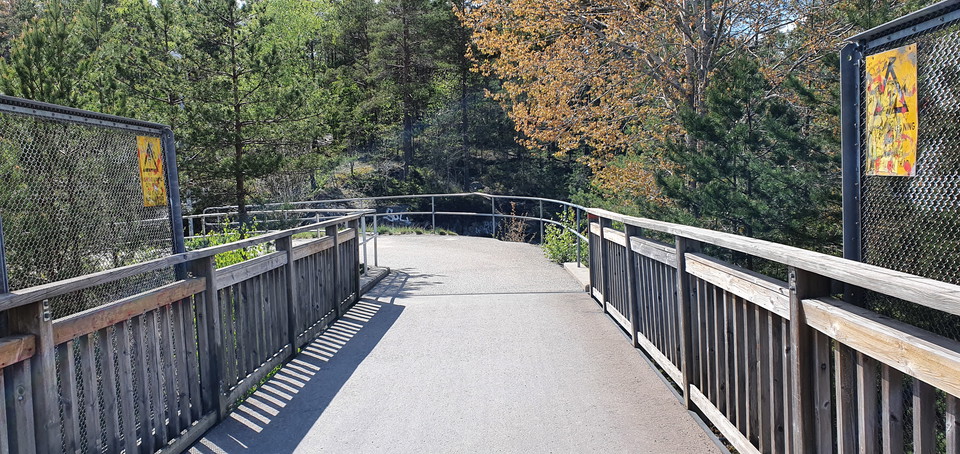 Gång- och cykelvägen svänger åt vänster. Räcken följer vägen. I bakgrunden syns skog som ligger i Rudans naturområde.