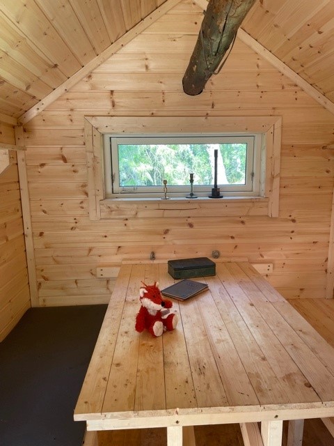 Inuti stuga, väggar och tak i naturfärgad träpanel, ett fönster rakt fram, två bänkar och ett bord.