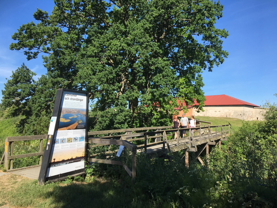 En borgruin med en våning och rött plåttak, i förgrunden en bro över vallgraven och tre personer som går.