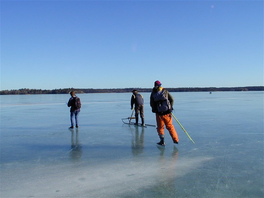Tre personer är på en istäckt sjö. Två åker långfärdsskridskor och en åker spark.