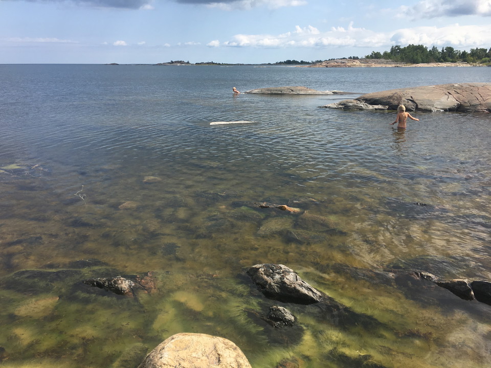 I en vik badar två personer. Det är långgrunt och genom vattnet ser man att botten är mycket stenig och ojämn.