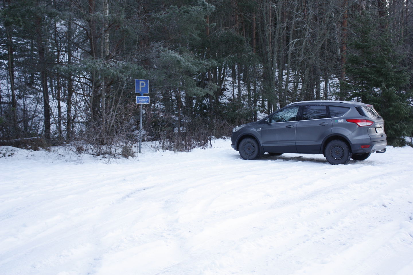 En bil står på en snötäckt parkeringsplats framför en skog. På parkeringen finns en parkeringsskylt.