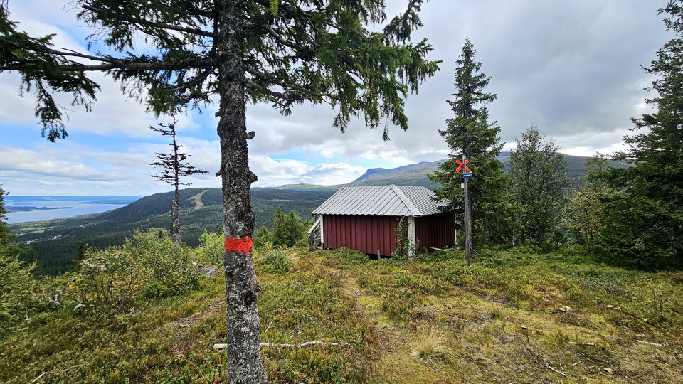Rött litet hus på toppen av ett berg. Bakom huset ser du milsvid utsikt över andra fjäll