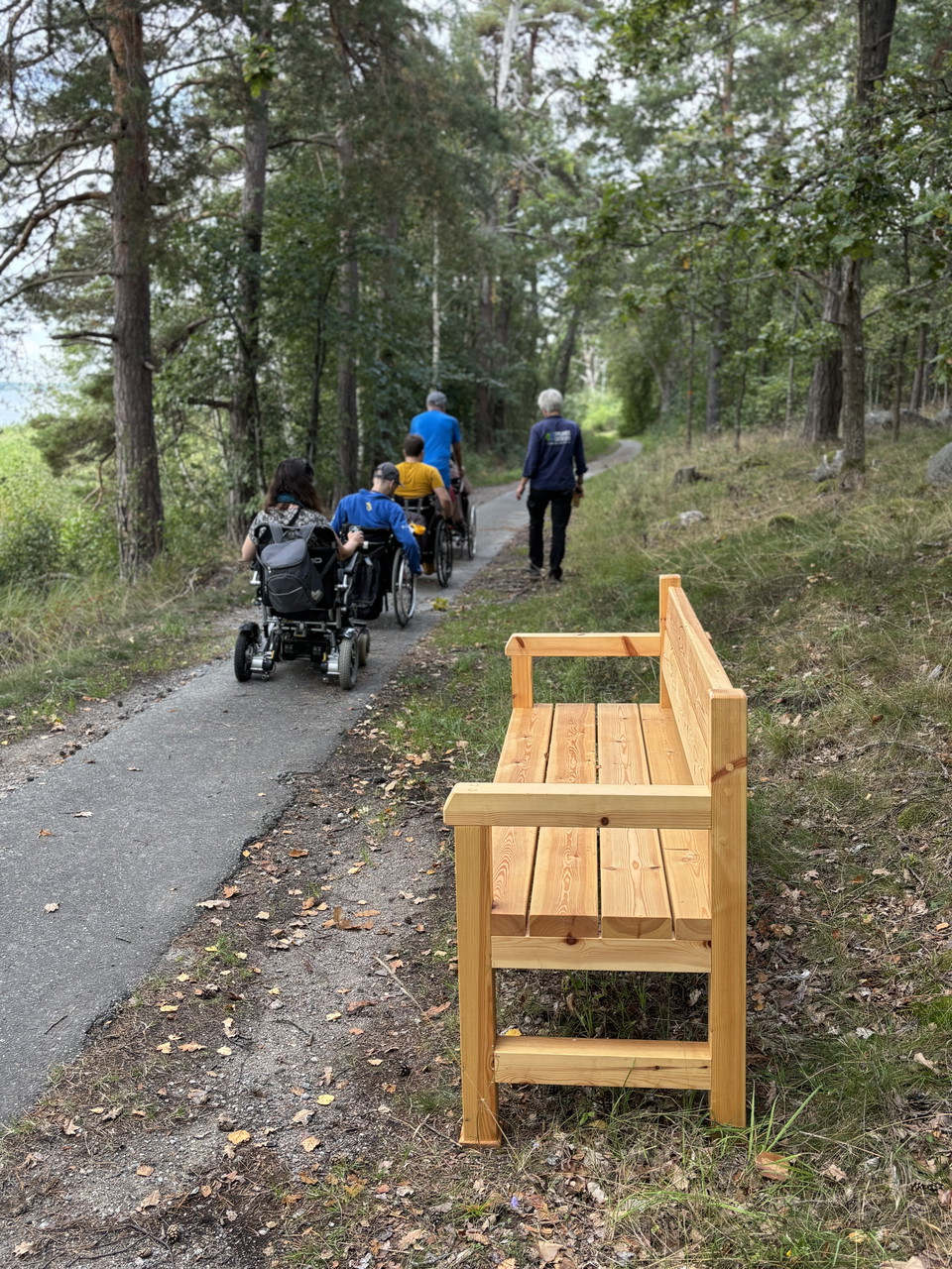 En träsoffa står längs en asfalterad gångväg. Några personer, varav flera i rullstol går längs gångvägen.