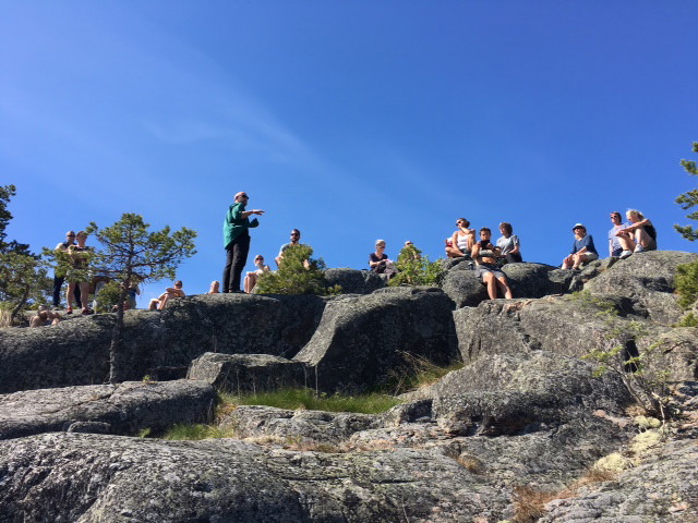 En stor grupp människor sitter på ojämna stenblock. Framför gruppen står en person och pekar åt höger.