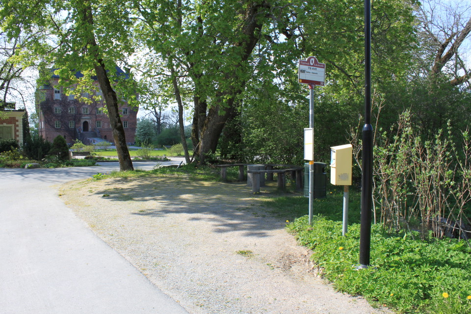 Vid en bilväg finns en busshållsplats och en brevlåda. I närheten står några sittbänkar och i bakgrunden står ett slott.