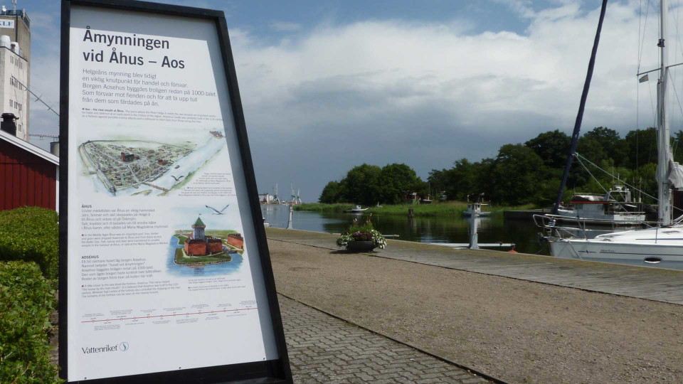 En informationstavla i form av ett segel i förgrunden. bakom skymtar Helgeåns utlopp i havet.