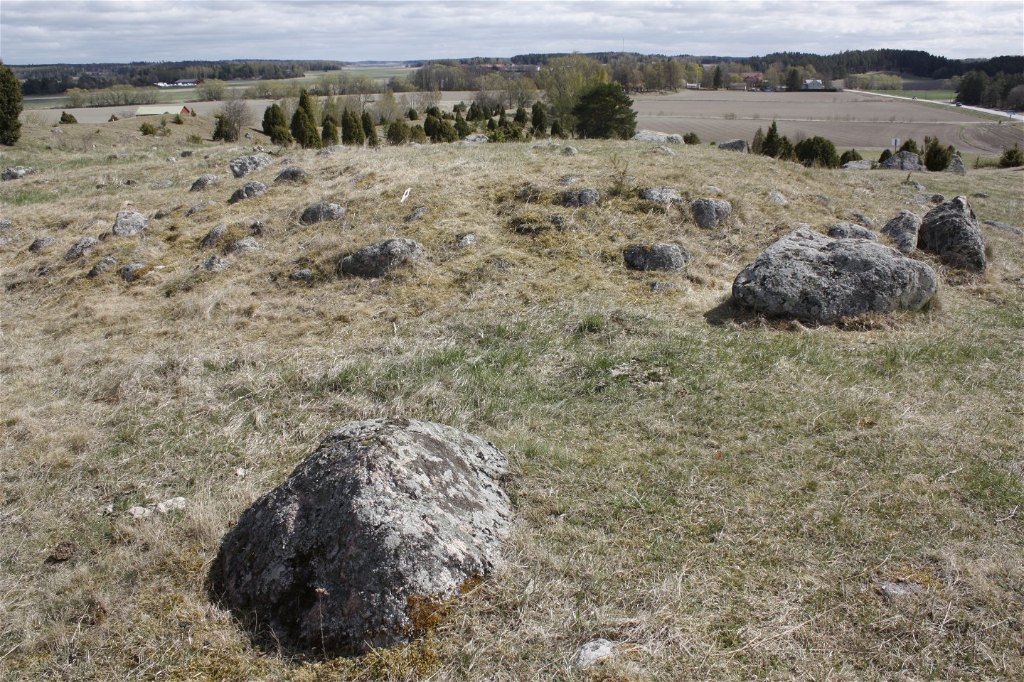 Flera större stenar ligger längs en sluttande ås. Nedanför finns jordbrukslandskap, gårdar och skog.