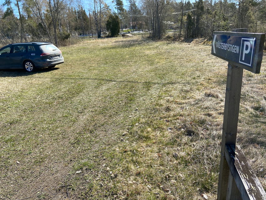 En bil står parkerad på en öppen gräsyta. Vid platsen finns en parkeringsskylt och i bakgrunden skymtar hus fram mellan träden.