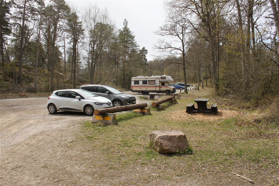 Flera bilar och en husbil står på en avlång grusad parkeringsplats. Framför parkeringen står ett bänkbord på en gräsplätt.