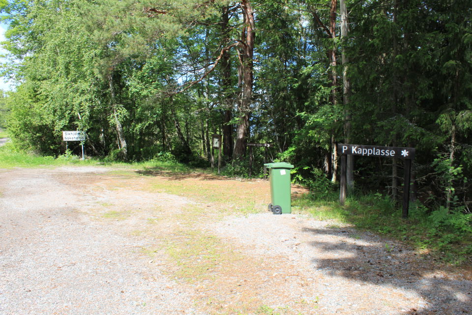 En grusad parkeringsplats med skog framför. På parkeringen står en soptunna, informationstavla och en skylt med texten Kapplasse.