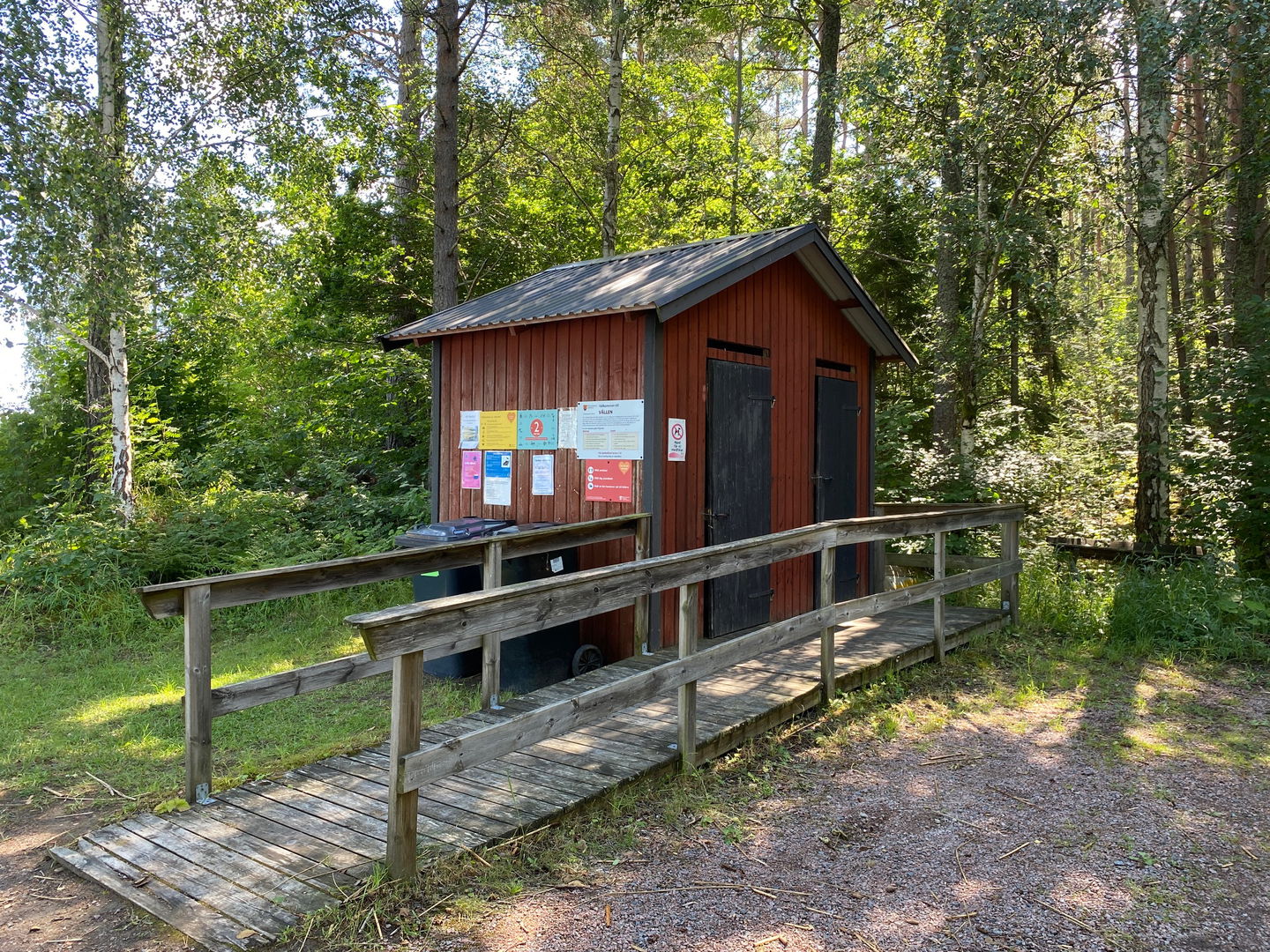 En ramp med handräcken leder fram till en torrtoalett med två dörrar. På sidan av toaletten finns informationstavla. Bakom toaletten är det skog.