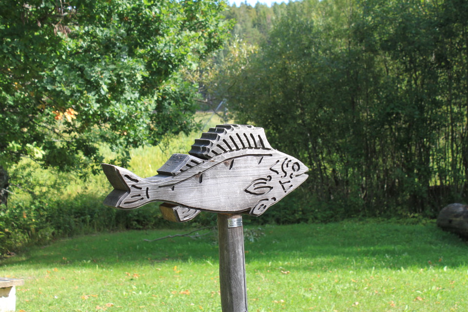 På en plan gräsyta står en trästolpe med en träskulptur av en fisk monterad på toppen. Runtomkring finns många träd.