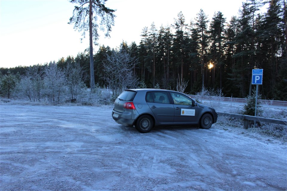 En bil står på en snötäckt parkerigsplats med en parkeringsskylt. I närheten av parkeringen går en bilväg.